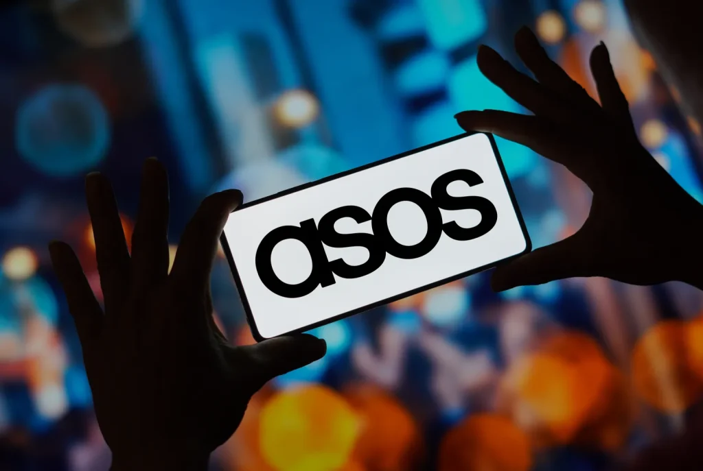 Hands holding a smartphone displaying the Asos logo, with blurred colorful lights in the background.