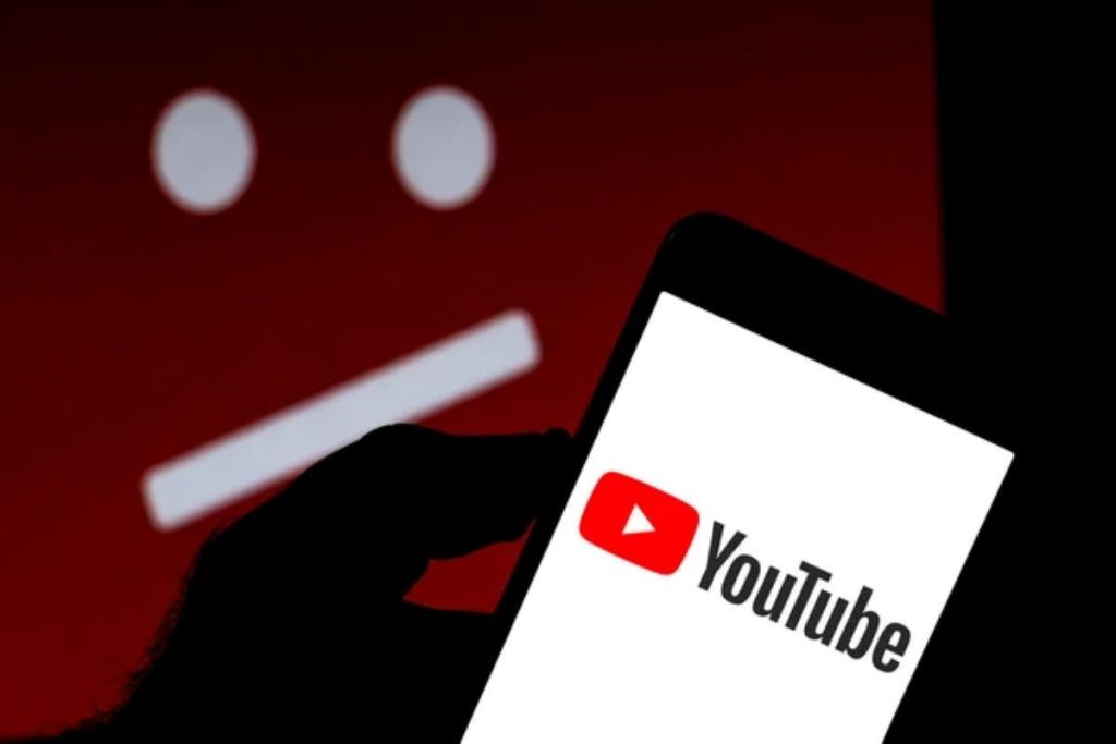 A hand holding a smartphone showing the YouTube logo on the screen, with a blurred red background featuring a frowning face icon symbolizing dissatisfaction.