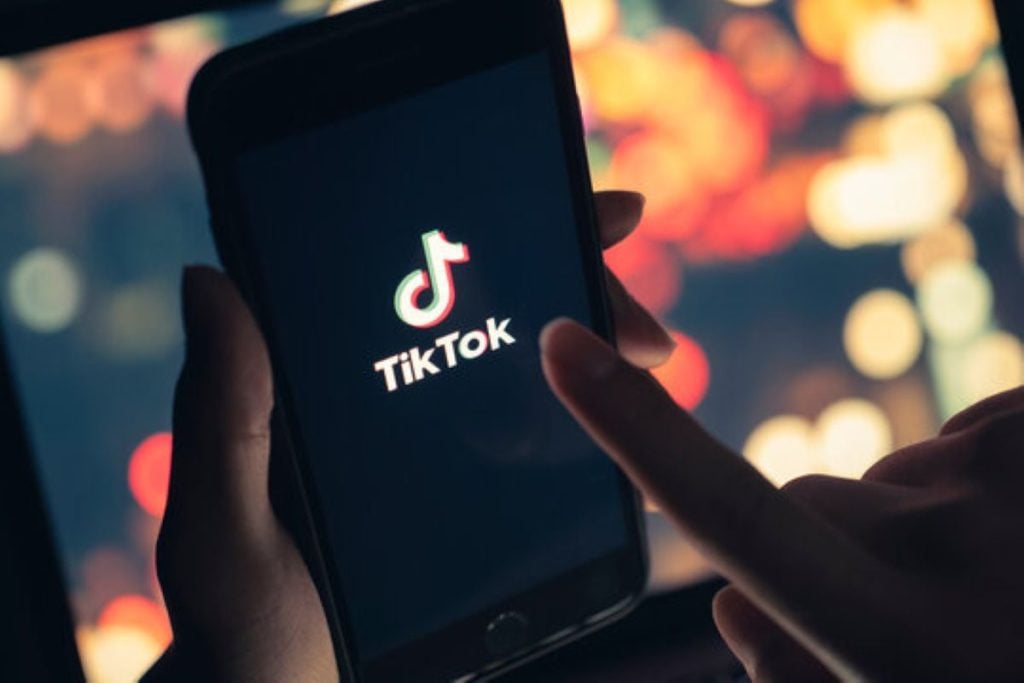 A person holding a smartphone displaying the TikTok logo on a dark screen with colorful blurred lights in the background.