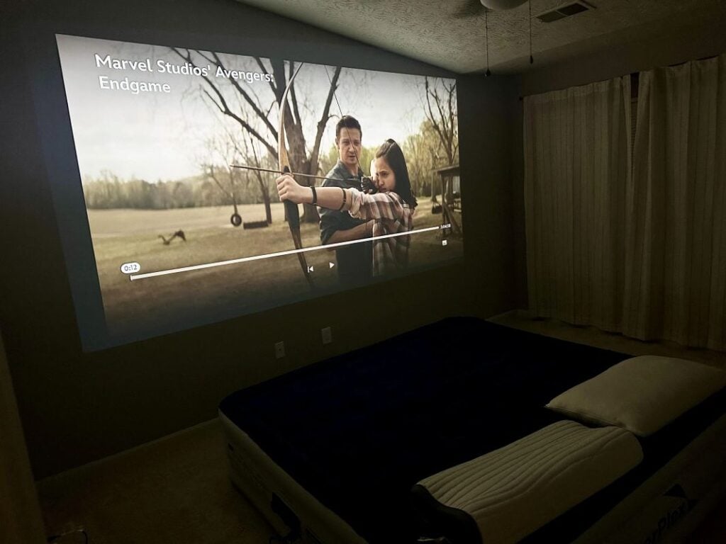 Dark bedroom with a large movie projected directly onto the wall above the bed for a cozy home theater setup.