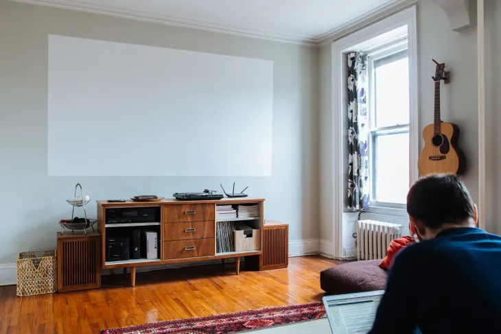 Bright apartment living room with a blank wall projector screen above a wooden media console and a person sitting nearby.