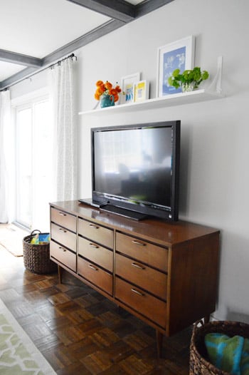 White floating shelf above a TV with small framed prints, flowers, and greenery over a wood dresser media console.