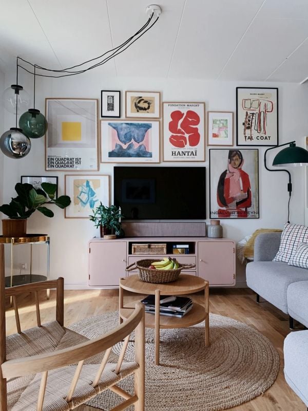 Gallery wall of colorful framed art arranged around a wall-mounted TV above a pale pink media console in a cozy living room.