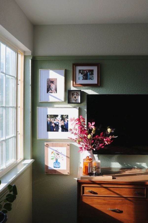 Green TV accent wall with adhesive-style framed photos and artwork placed beside a wall-mounted television near a sunny window.