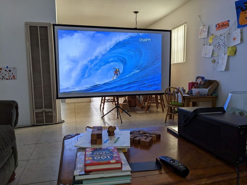 Portable projector screen on a tripod showing an ocean surfing video in a living room with a projector on the coffee table.