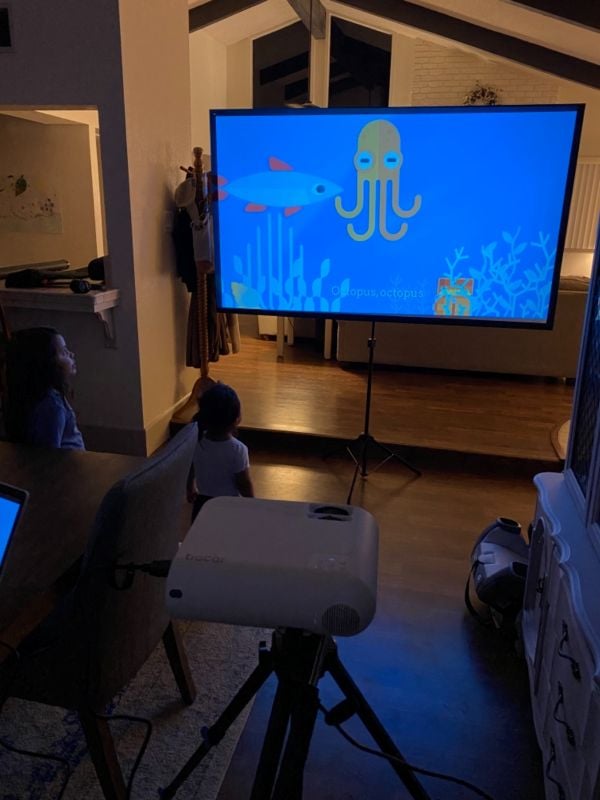 Portable projector screen on a tripod showing a children’s cartoon in a dim living room while kids watch from the couch.
