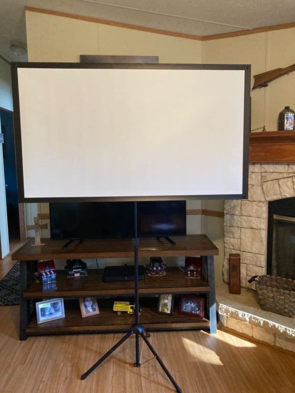Blank portable projector screen on a tripod stand placed in front of a TV console and fireplace.