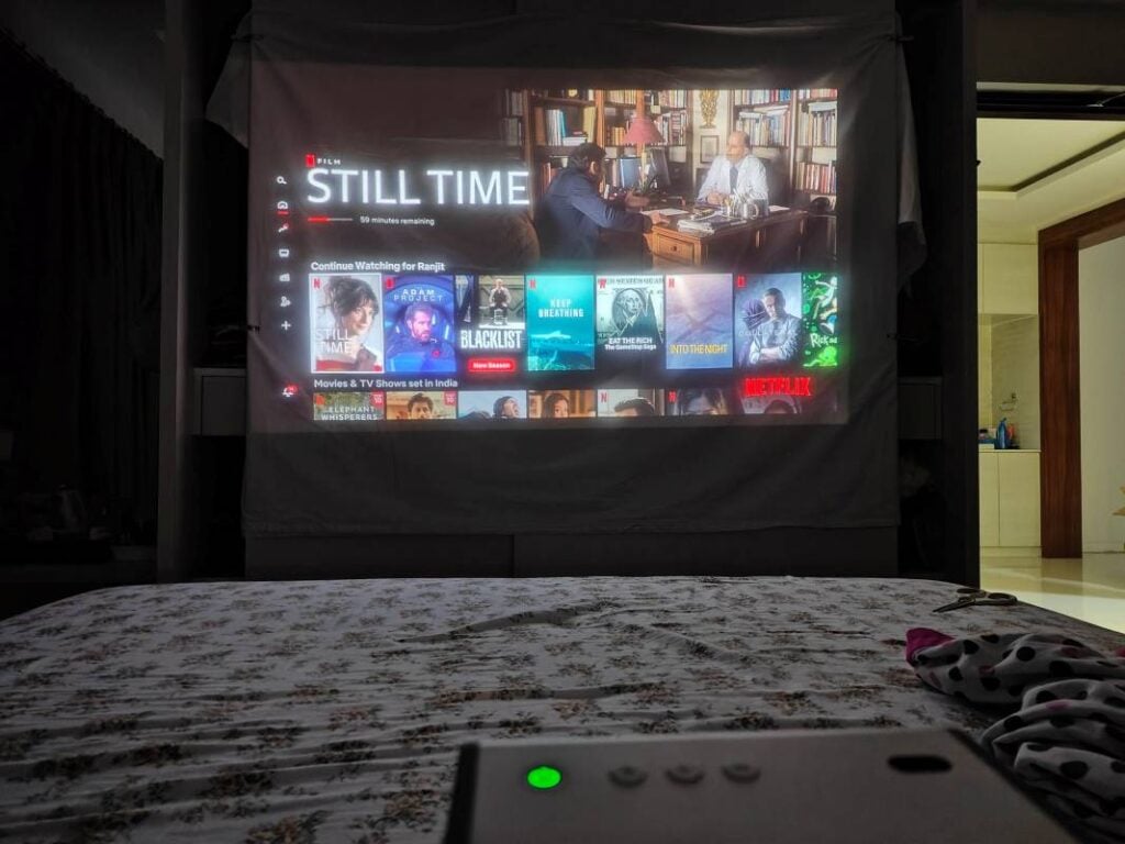 Netflix home screen projected onto a large white sheet in a dark bedroom with a remote in the foreground.