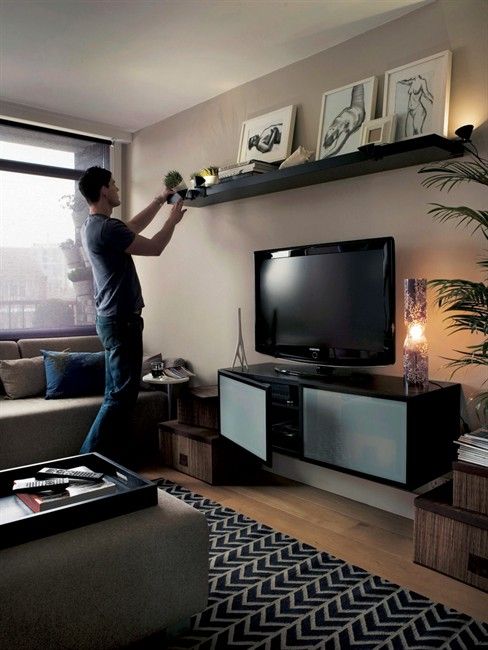 Person decorating a long floating shelf above a TV with framed artwork, plants, and accent lighting in a modern living room.