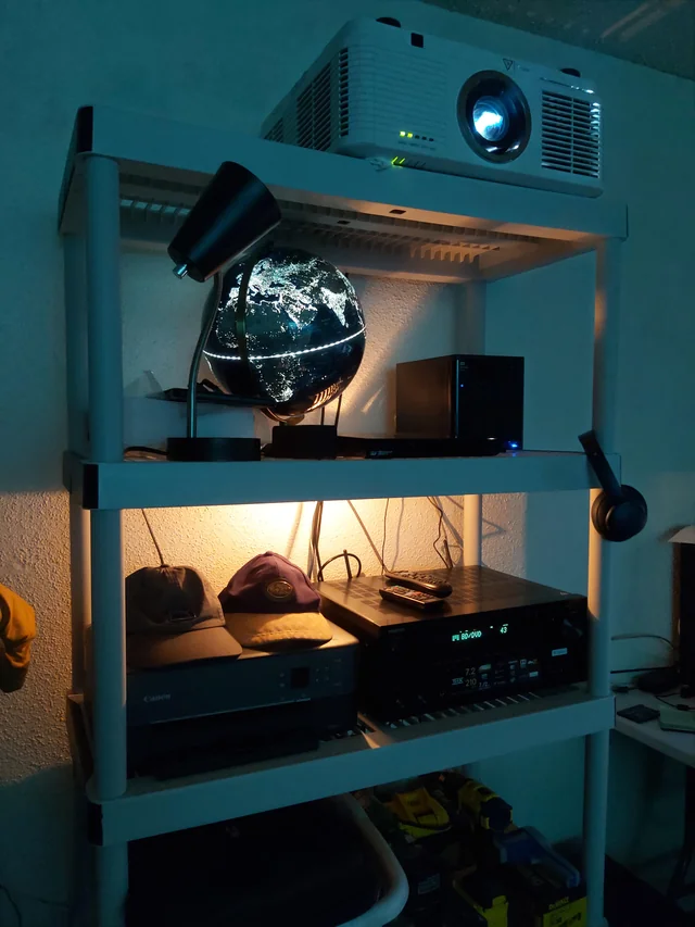 Projector placed on a white shelving unit above audio equipment and a glowing globe lamp.