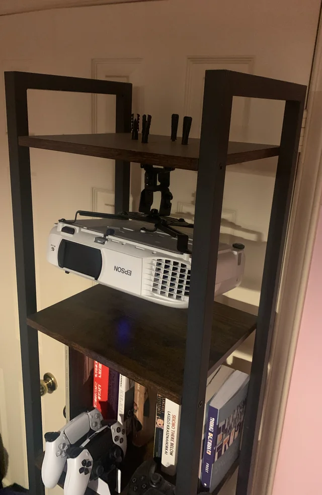 Epson projector set on a bookshelf shelf with gaming controllers and books stored underneath.