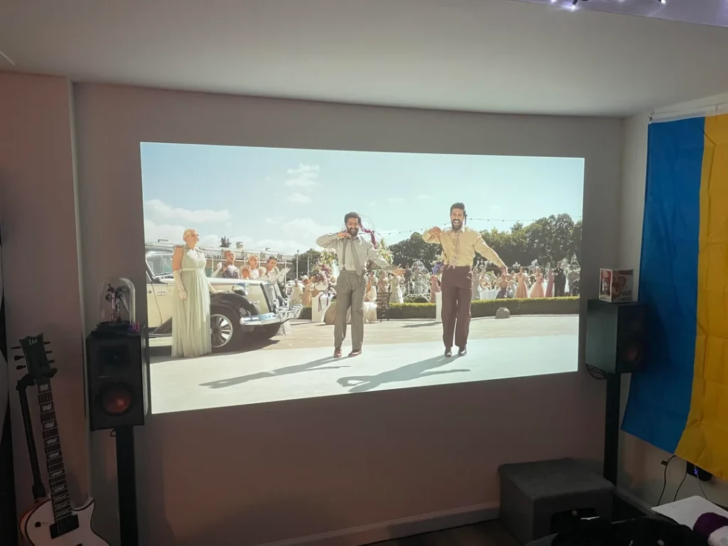 Large movie projection displayed directly on a blank wall in a room with speakers, a guitar, and a wall flag.