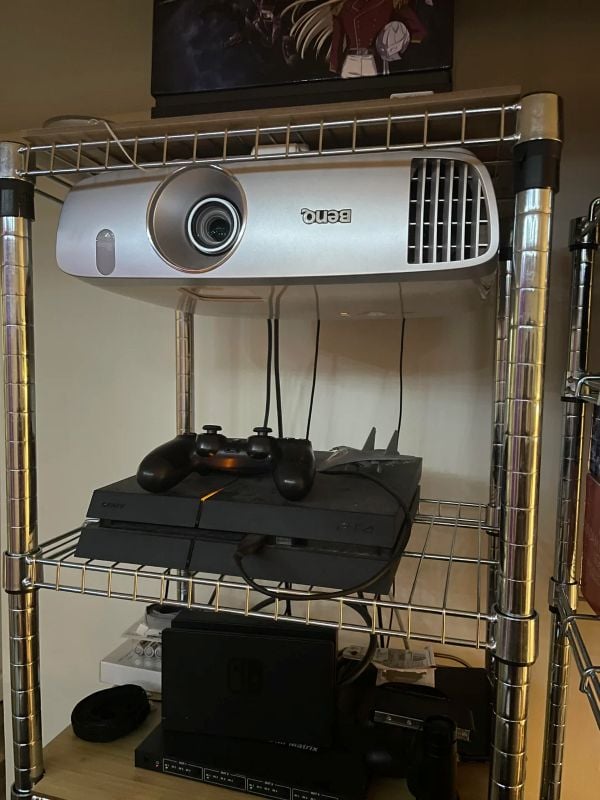 BenQ projector mounted upside down under a metal shelving rack above gaming consoles.