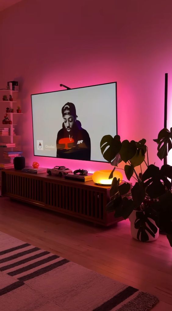 Modern living room TV wall with bright pink ambient LED lighting, a low wood console, and large houseplants.