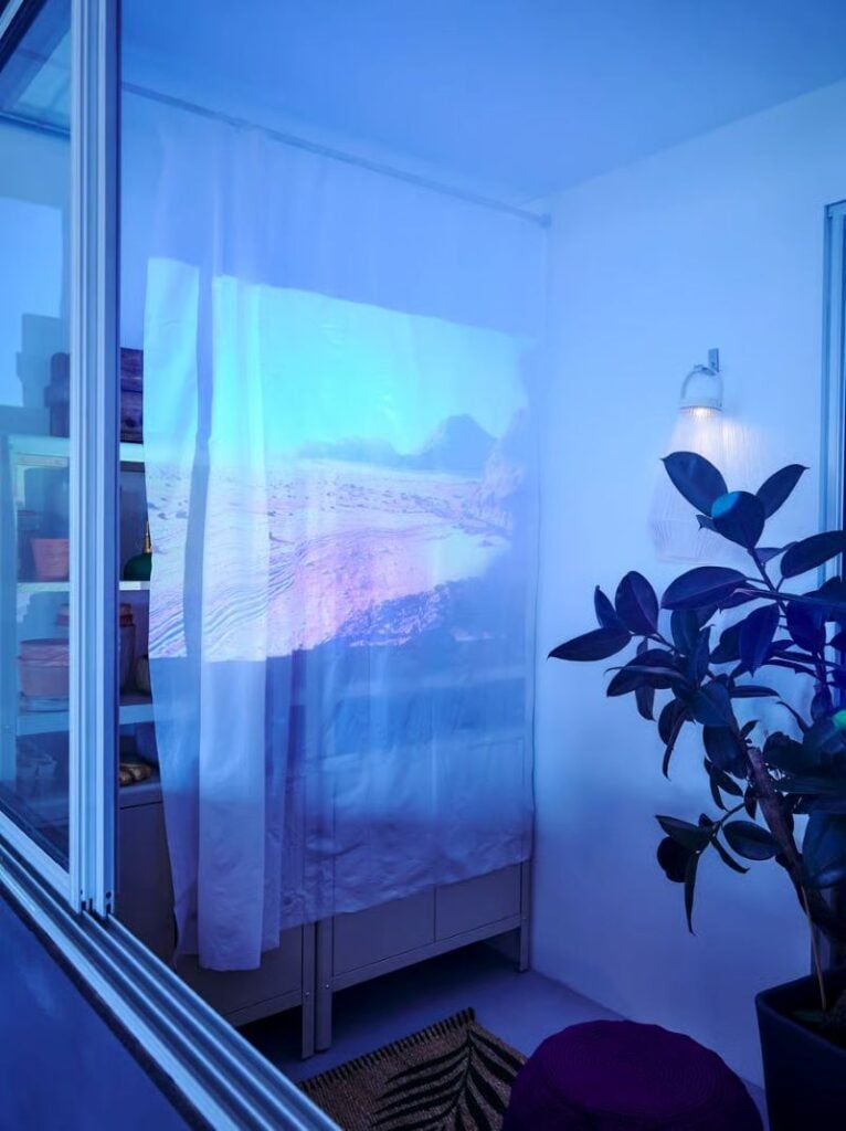 A small projector casts a soft beach scene onto a white curtain inside a blue-lit room, creating a cozy indoor movie setup.