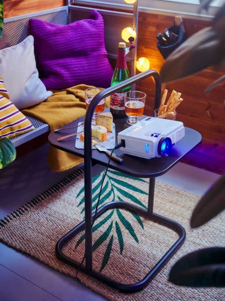 A white mini projector sits on a coffee table beside drinks and snacks in a colorful lounge-style living room.
