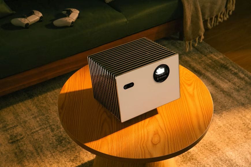 A compact rectangular projector rests on a round wooden coffee table in a dim living room with warm lighting.