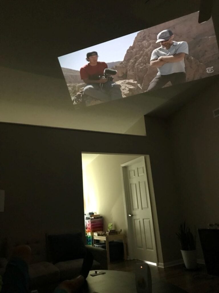 A movie scene is projected high onto a wall near the ceiling in a dark living room with an open doorway below.