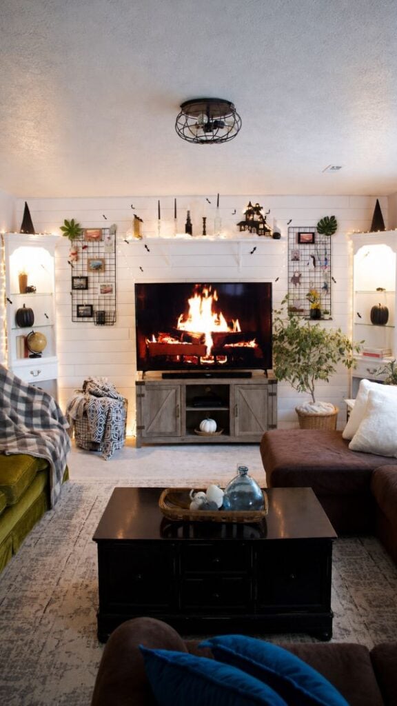Cozy living room TV setup with a fireplace video on screen, black grid wall decor, shelves, and seasonal accents.