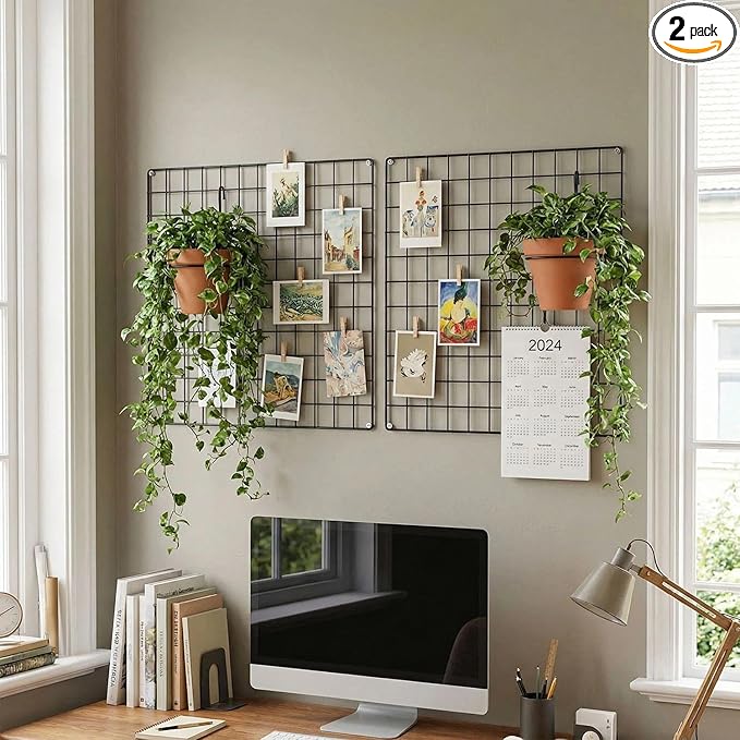 Home office wall with black wire grid panels, clipped artwork, hanging plants, and a desktop monitor below.