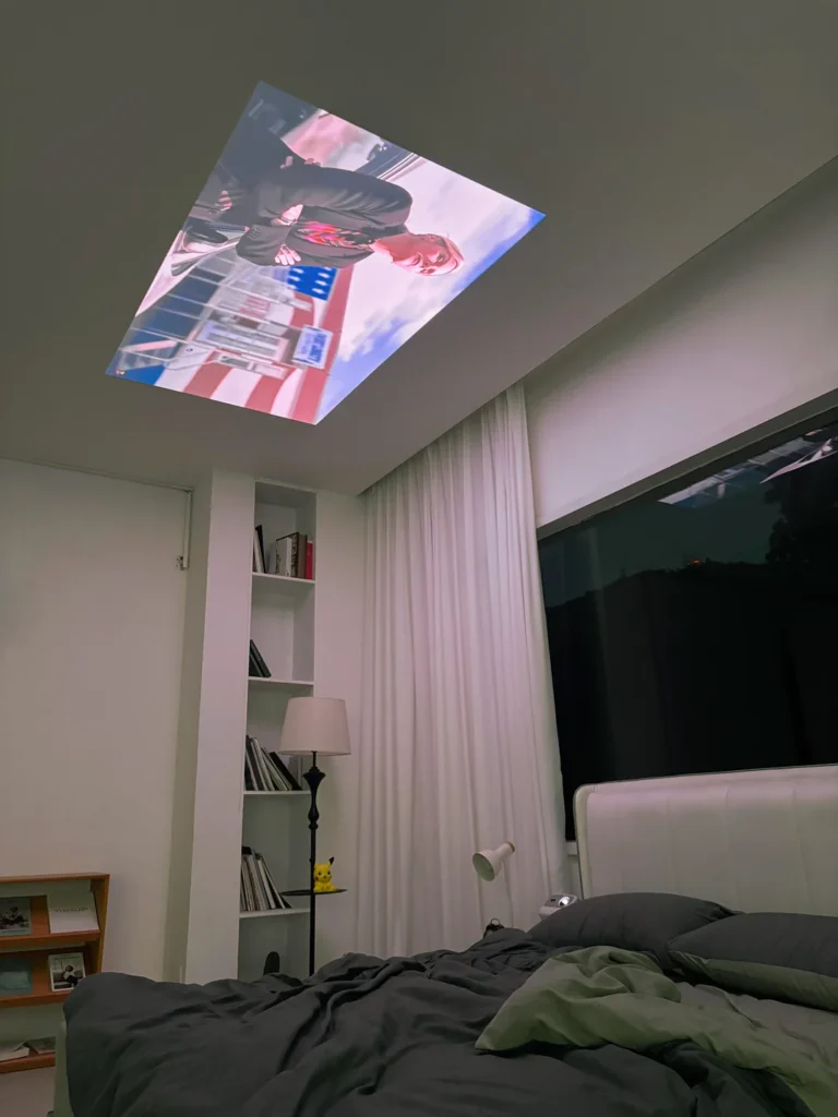 A bedroom projector setup shows a movie projected onto the ceiling above a bed at night.