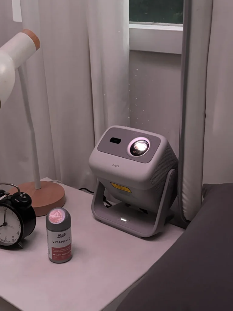 A compact projector sits on a bedside table beside a lamp, clock, and vitamin bottle, aimed upward for ceiling viewing.