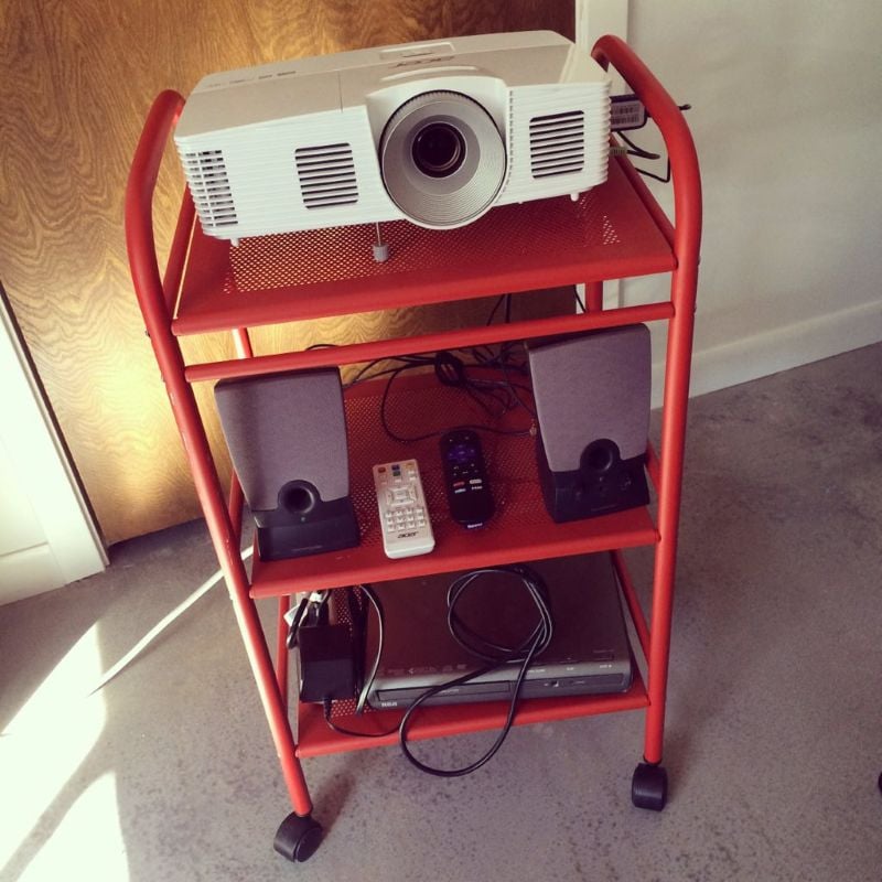 A white projector sits on the top shelf of a red rolling cart, with speakers, remotes, and media equipment stored on the shelves below.