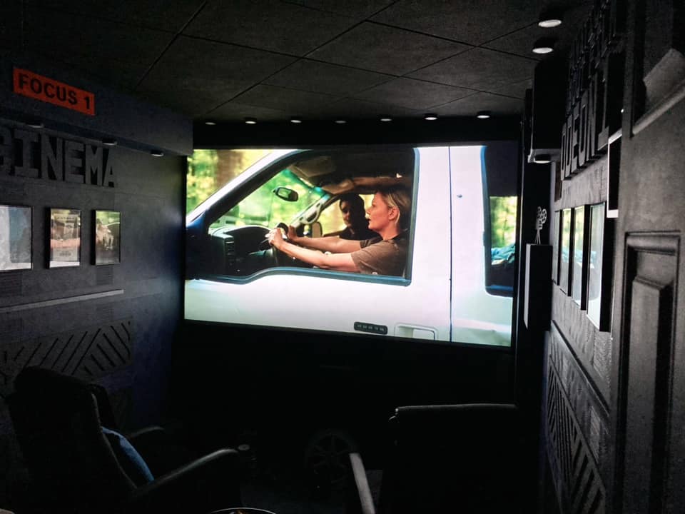 A home theater room shows a large movie projected onto a wall screen in a dark cinema-style space.