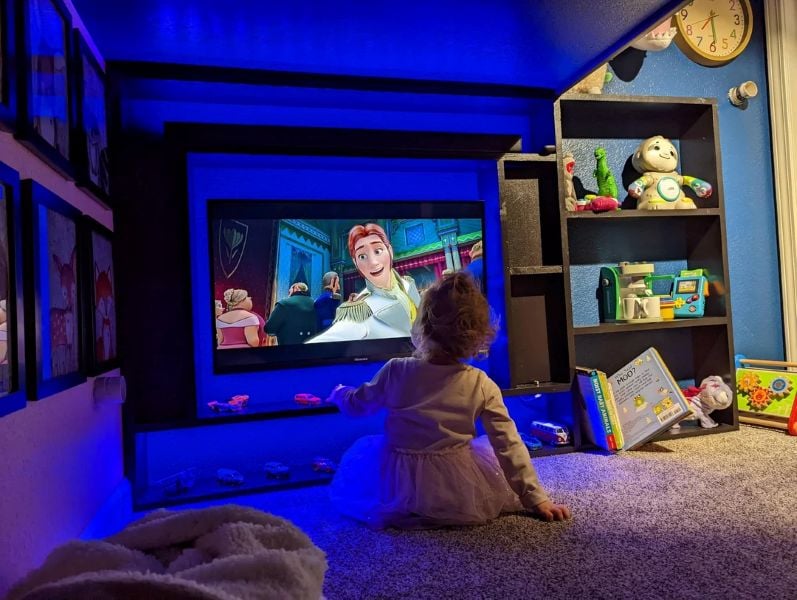 A toddler sits on the carpet watching an animated movie projected onto a screen in a cozy blue-lit playroom.