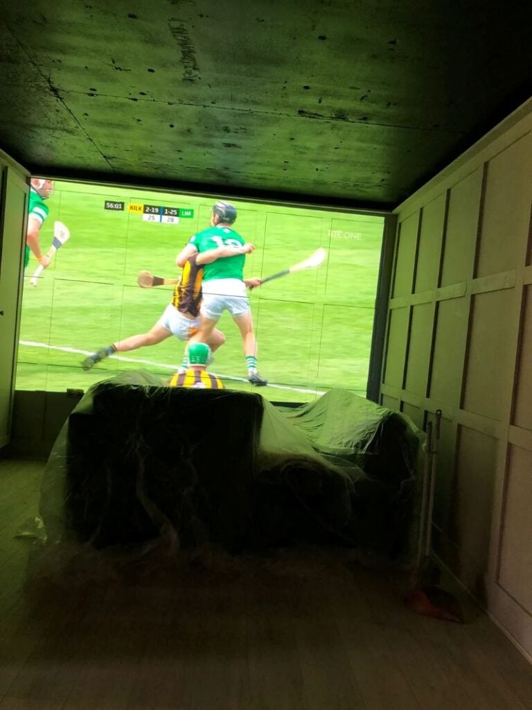 A projector casts a sports game onto a large wall inside a dim closet or garage-style room with covered furniture.