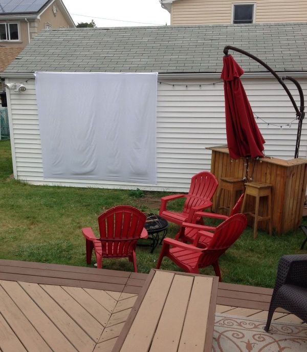 A backyard patio setup uses a white sheet hung on an exterior wall as an outdoor projector screen.