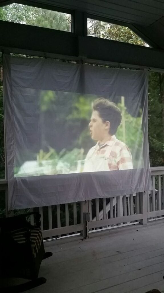 A movie is projected onto a white sheet hanging on a screened porch, creating a simple outdoor theater setup.