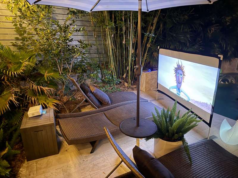 An outdoor patio projector setup shows a movie playing on a freestanding screen beside lounge chairs and garden plants.
