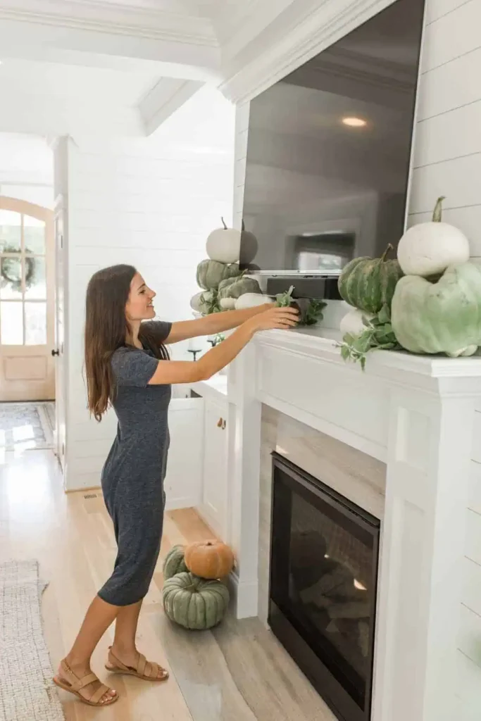Woman decorating a fireplace mantel under a mounted TV with pumpkins and greenery for a seasonal fall display.