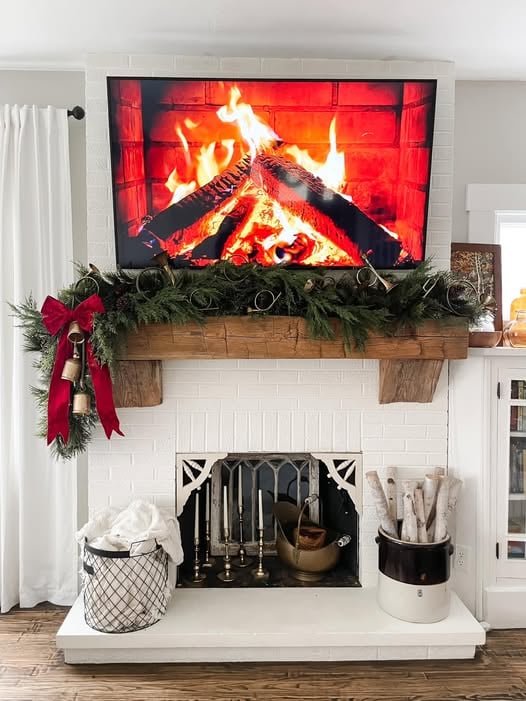 Fireplace mantel with a TV showing a fire scene, decorated with evergreen garland, bells, and holiday accents.