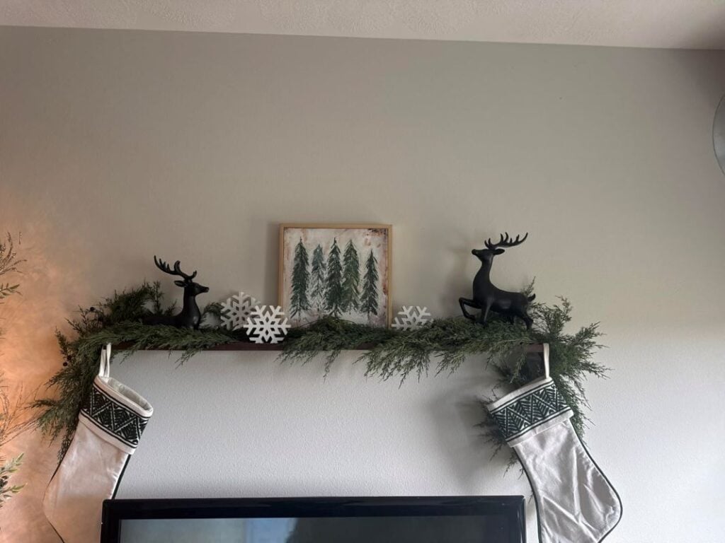 Holiday floating shelf above a TV styled with greenery, stockings, snowflakes, framed art, and black reindeer.