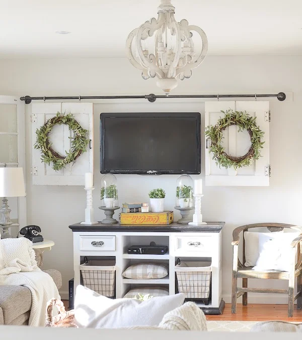TV wall with a mounted television between two white shutter-style panels, a curtain rod above, and green wreaths used as decorative accents.
