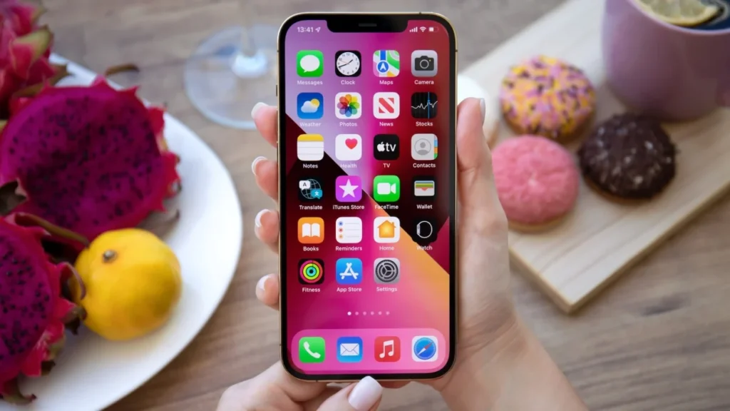 Hand holding an iPhone displaying a home screen filled with preinstalled apps, with colorful donuts and fruit on a table in the background.
