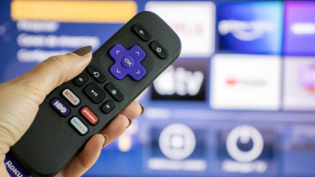 Hand holding a Roku remote in front of a blurred TV screen with streaming app tiles.