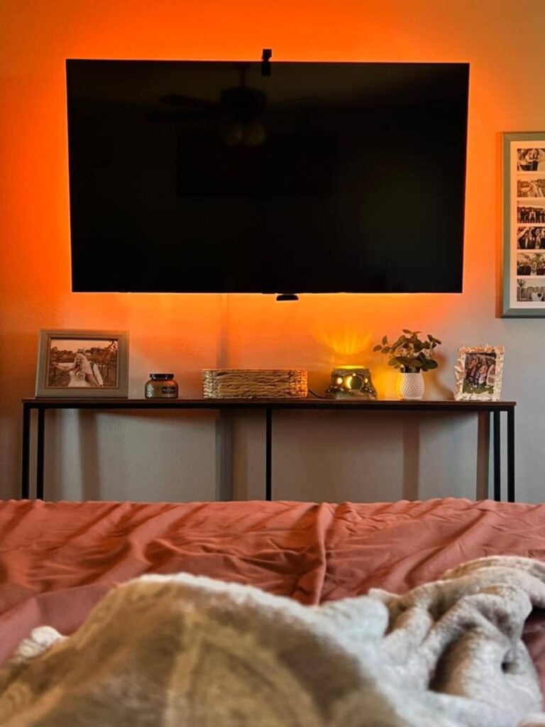Wall-mounted TV above a console table with warm backlighting and a visible cord beneath the screen.