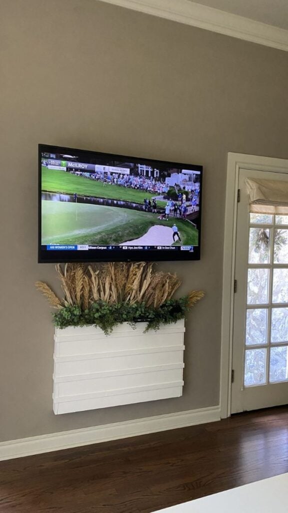 Wall-mounted TV above a white decorative wall planter filled with dried grasses, greenery, and hidden cable storage.