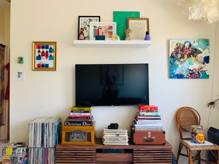 TV displayed on a wooden media console surrounded by stacked books, records, artwork, and a wall shelf for a colorful eclectic setup.